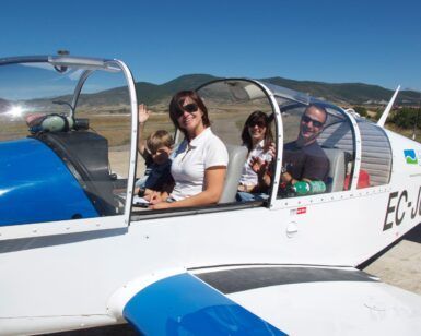 Una familia de cuatro personas disfruta de su vuelo en Avioneta sobre el Parque de Ordesa, sonriendo y saludando dentro de una pequeña avioneta en un día soleado con las pintorescas colinas del parque de fondo. - ForTwo - Planes para parejas