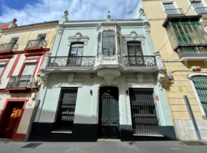 Un edificio histórico con un balcón ornamentado y una fachada intrincada está situado entre edificios beige y amarillos bajo un cielo azul, ofreciendo una visión del Hotel con encanto en el centro de Badajoz. - ForTwo - Planes para parejas