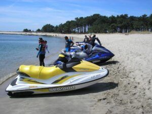 En la playa de arena junto a un pinar y bajo un cielo azul claro, la gente con traje de neopreno se prepara para disfrutar de "Alquila tu moto de agua en Vilagarcía de Arousa". - ForTwo - Planes para parejas