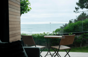 Dos sillas tejidas y una mesa en el patio del Apartahotel en Playa de Valcovo ofrecen vistas al océano nublado enmarcado por exuberante vegetación y árboles. - ForTwo - Planes para parejas