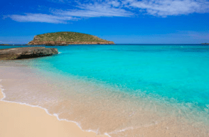 Una playa de agua turquesa clara, orilla de arena y una pequeña isla bajo un cielo azul crean el escenario perfecto para "Aventura en lancha: Playas y cuevas de Ibiza. - ForTwo - Planes para parejas