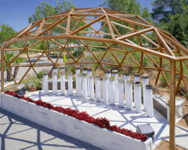 En BIBO PARK Ibiza: Naturaleza y biotecnología, una elegante cúpula de madera abierta con columnas cilíndricas blancas se alza entre vibrantes flores rojas bajo el cielo azul claro. - ForTwo - Planes para parejas