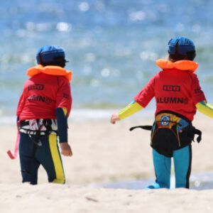 Dos personas con trajes de neopreno y chalecos salvavidas se encuentran en la playa de arena de Pontevedra, con el océano detrás de ellos, listos para un emocionante Bautismo de Kitesurf en Pontevedra. - ForTwo - Planes para parejas