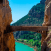 El estrecho puente de piedra, muy parecido al aventurero Caminito del Rey: Aventura desde Málaga, se extiende elegantemente entre acantilados sobre un río turquesa, rodeado de exuberante vegetación bajo un cielo azul brillante. - ForTwo - Planes para parejas