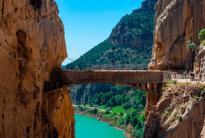 El estrecho puente de piedra, muy parecido al aventurero Caminito del Rey: Aventura desde Málaga, se extiende elegantemente entre acantilados sobre un río turquesa, rodeado de exuberante vegetación bajo un cielo azul brillante. - ForTwo - Planes para parejas