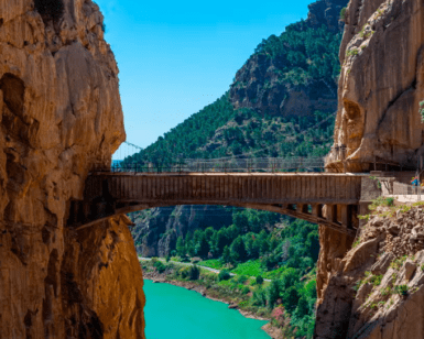 El estrecho puente de piedra, muy parecido al aventurero Caminito del Rey: Aventura desde Málaga, se extiende elegantemente entre acantilados sobre un río turquesa, rodeado de exuberante vegetación bajo un cielo azul brillante. - ForTwo - Planes para parejas