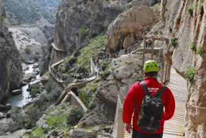 Una persona con una chaqueta roja recorre el estrecho sendero de montaña del Caminito del Rey, disfrutando del "Caminito del Rey: Aventura desde Málaga", rodeado de impresionantes paisajes rocosos y compartiendo el emocionante viaje con otros. - ForTwo - Planes para parejas