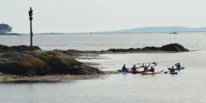 Un grupo de kayakistas comienza un emocionante viaje del Camino de Santiago en Kayak a lo largo de costas rocosas en una vasta y tranquila masa de agua bajo un cielo brumoso. - ForTwo - Planes para parejas