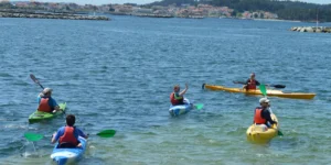 Cuatro personas disfrutan de un paseo en kayak bajo el sol en tranquilas aguas con el pintoresco pueblo de Santiago de fondo, mostrando la experiencia del "Camino de Santiago en Kayak". - ForTwo - Planes para parejas