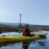 Una persona se desliza en un kayak amarillo por aguas tranquilas junto a una orilla rocosa, donde una cruz y un árbol hacen centinela, capturando el sereno recorrido del Camino de Santiago en Kayak, combinando aventura con reflexión espiritual. - ForTwo - Planes para parejas