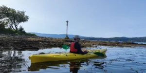 Una persona se desliza en un kayak amarillo por aguas tranquilas junto a una orilla rocosa, donde una cruz y un árbol hacen centinela, capturando el sereno recorrido del Camino de Santiago en Kayak, combinando aventura con reflexión espiritual. - ForTwo - Planes para parejas