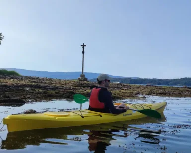 Una persona se desliza en un kayak amarillo por aguas tranquilas junto a una orilla rocosa, donde una cruz y un árbol hacen centinela, capturando el sereno recorrido del Camino de Santiago en Kayak, combinando aventura con reflexión espiritual. - ForTwo - Planes para parejas