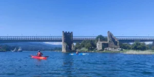 Dos kayakistas reman bajo un puente entre antiguas ruinas de piedra en un día soleado, evocando el sereno viaje que ofrece el Camino de Santiago en Kayak. - ForTwo - Planes para parejas