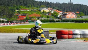 Con casco puesto, un corredor acelera en un kart en el Kartódromo Valga con montones de neumáticos y casas con encanto cerca, capturando la emoción de "Carreras de karts en Kartódromo Valga en Pontevedra. - ForTwo - Planes para parejas