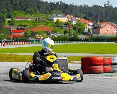 Con casco puesto, un corredor acelera en un kart en el Kartódromo Valga con montones de neumáticos y casas con encanto cerca, capturando la emoción de "Carreras de karts en Kartódromo Valga en Pontevedra. - ForTwo - Planes para parejas