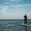 Vestido con un traje de neopreno, alguien saborea el paddleboarding en el mar tranquilo bajo un cielo azul claro, capturando la esencia de las Clases de Paddle Surf en O Grove. - ForTwo - Planes para parejas