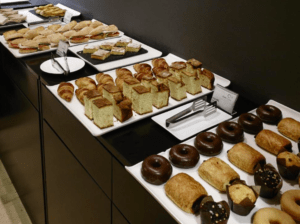 La Escapada romántica en el Hotel Carris Marineda cuenta con una mesa de buffet bellamente dispuesta con una variedad de pasteles, sándwiches, donas, muffins y pasteles en elegantes bandejas blancas colocadas contra una pared oscura. - ForTwo - Planes para parejas