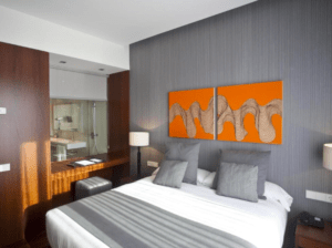 Experimente la elegancia moderna en el Hotel Carris Marineda con una lujosa cama doble, acogedoras almohadas grises y vibrantes obras de arte en naranja. Ideal para una escapada romántica en el Hotel Carris Marineda, disfrute de la vista única del baño, que combina diseño contemporáneo con un encanto romántico. - ForTwo - Planes para parejas