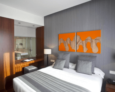Experimente la elegancia moderna en el Hotel Carris Marineda con una lujosa cama doble, acogedoras almohadas grises y vibrantes obras de arte en naranja. Ideal para una escapada romántica en el Hotel Carris Marineda, disfrute de la vista única del baño, que combina diseño contemporáneo con un encanto romántico. - ForTwo - Planes para parejas