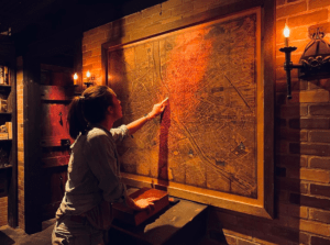 Una persona señala un gran mapa antiguo en una pared de ladrillos con poca luz, invitándote a entrar en "Escape Room Operación Bunker: ¡Desafía tu ingenio!. - ForTwo - Planes para parejas