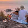 Mientras la pareja se sienta en un acantilado al atardecer con su picnic preparado, recuerdan su día de "Excursión 4x4, senderismo y barco en Ibiza", saboreando la tranquilidad y esperando avistar un barco lejano en el horizonte. - ForTwo - Planes para parejas