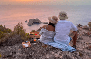 Mientras la pareja se sienta en un acantilado al atardecer con su picnic preparado, recuerdan su día de "Excursión 4x4, senderismo y barco en Ibiza", saboreando la tranquilidad y esperando avistar un barco lejano en el horizonte. - ForTwo - Planes para parejas