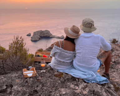 Mientras la pareja se sienta en un acantilado al atardecer con su picnic preparado, recuerdan su día de "Excursión 4x4, senderismo y barco en Ibiza", saboreando la tranquilidad y esperando avistar un barco lejano en el horizonte. - ForTwo - Planes para parejas
