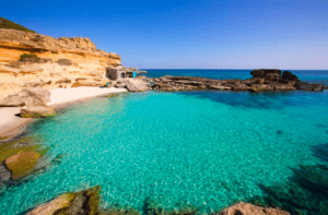 Experimenta Excursión a Formentera: Un día en el paraíso, donde aguas cristalinas de color turquesa se encuentran con una cala rocosa con una pequeña playa de arena bajo un cielo azul brillante, capturando la verdadera esencia de Formentera. - ForTwo - Planes para parejas