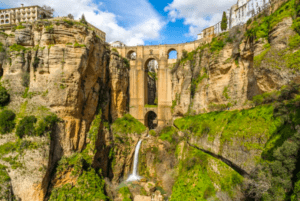 Bajo el cielo azul de Málaga, la Excursión a Ronda y Setenil desde Málaga revela el encantador puente de piedra de Ronda que se extiende majestuosamente sobre un profundo desfiladero con exuberante vegetación y una cascada debajo, creando una escena verdaderamente cautivadora. - ForTwo - Planes para parejas