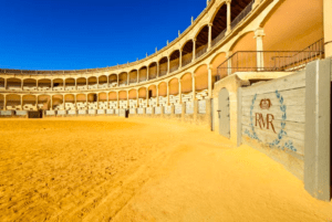 Vista interior de una histórica plaza de toros de Ronda iluminada por el sol con asientos curvos y suelo de arena, parte de la "Excursión a Ronda y Setenil desde Málaga. - ForTwo - Planes para parejas