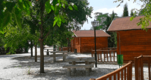 Cabañas de madera y mesas de picnic descansan bajo árboles soleados en un tranquilo parque con senderos de grava que serpentean junto a una farola. Ideal para una escapada de fin de semana en el Embalse de Negratín, rodeado de una serena belleza. - ForTwo - Planes para parejas