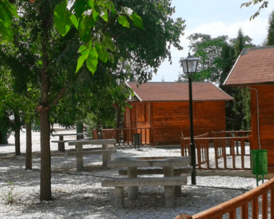 Cabañas de madera y mesas de picnic descansan bajo árboles soleados en un tranquilo parque con senderos de grava que serpentean junto a una farola. Ideal para una escapada de fin de semana en el Embalse de Negratín, rodeado de una serena belleza. - ForTwo - Planes para parejas