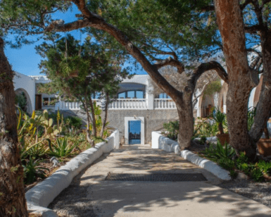Un camino arbolado conduce al Hostal La Torre Ibiza Cap Negre Ibiza, un edificio blanco adornado con arcos, que ofrece impresionantes vistas al mar que recuerdan el encanto de Ibiza. - ForTwo - Planes para parejas