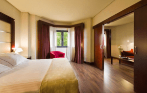 Disfrute de la modernidad y el bienestar en el Hotel Badajoz Center en una espaciosa habitación con una cama King, suelo de madera y puertas corredizas que conducen a una zona de estar con una silla roja. - ForTwo - Planes para parejas