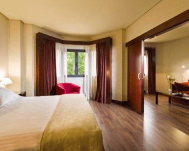 Disfrute de la modernidad y el bienestar en el Hotel Badajoz Center en una espaciosa habitación con una cama King, suelo de madera y puertas corredizas que conducen a una zona de estar con una silla roja. - ForTwo - Planes para parejas