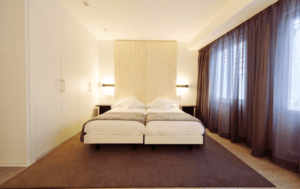 Experimente la tranquila elegancia del dormitorio minimalista del Hotel Lois en A Coruña con dos camas individuales, una alfombra grande, cortinas marrones, iluminación suave en una pared con acento de madera: un santuario ideal para la relajación. - ForTwo - Planes para parejas