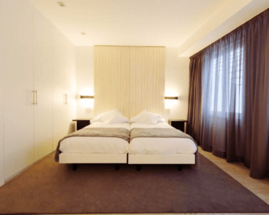 Experimente la tranquila elegancia del dormitorio minimalista del Hotel Lois en A Coruña con dos camas individuales, una alfombra grande, cortinas marrones, iluminación suave en una pared con acento de madera: un santuario ideal para la relajación. - ForTwo - Planes para parejas