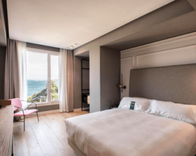 Alójese en el Hotel Riazor en La Coruña y disfrute de una moderna habitación con una cama grande, escritorio y TV. Cortinas transparentes enmarcan impresionantes vistas al mar para una experiencia encantadora desde su ventana. - ForTwo - Planes para parejas