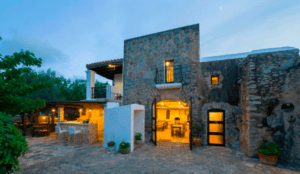 Un edificio rústico de piedra con un interior iluminado, asientos al aire libre y árboles bajo un cielo oscuro, perfecto para una escapada idílica en el Hotel Rural: Escapada al corazón de Ibiza. - ForTwo - Planes para parejas