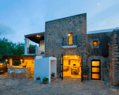 Un edificio rústico de piedra con un interior iluminado, asientos al aire libre y árboles bajo un cielo oscuro, perfecto para una escapada idílica en el Hotel Rural: Escapada al corazón de Ibiza. - ForTwo - Planes para parejas