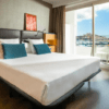 Alójese en el Hotel en Marina Botafoch Ibiza para disfrutar de una habitación moderna con una cama grande con almohadas azules, con una ubicación ideal para disfrutar de las vistas al mar y a la ciudad a través de una puerta corrediza de cristal. - ForTwo - Planes para parejas