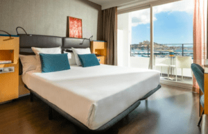 Alójese en el Hotel en Marina Botafoch Ibiza para disfrutar de una habitación moderna con una cama grande con almohadas azules, con una ubicación ideal para disfrutar de las vistas al mar y a la ciudad a través de una puerta corrediza de cristal. - ForTwo - Planes para parejas