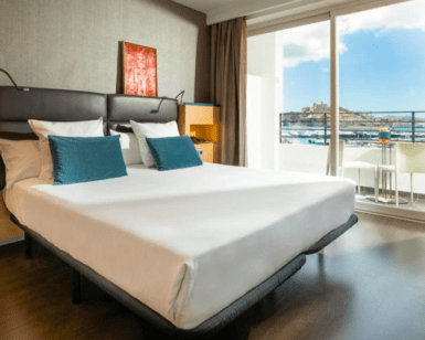 Alójese en el Hotel en Marina Botafoch Ibiza para disfrutar de una habitación moderna con una cama grande con almohadas azules, con una ubicación ideal para disfrutar de las vistas al mar y a la ciudad a través de una puerta corrediza de cristal. - ForTwo - Planes para parejas