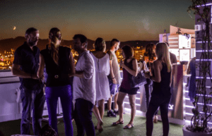 Al anochecer, la gente socializa en un bar en la azotea de Marina Botafoch, rodeado por el brillante horizonte iluminado por el Hotel en Marina Botafoch Ibiza y las luces de la ciudad, con una luna creciente en el fondo. - ForTwo - Planes para parejas