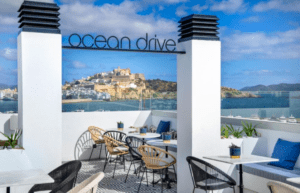 El Hotel en Marina Botafoch Ibiza cuenta con una terraza en la azotea con mesas y sillas blancas, que ofrece impresionantes vistas al mar y un cartel de Ocean Drive bajo un cielo azul. Experimente el vibrante ambiente del emblemático Marina Botafoch de Ibiza. - ForTwo - Planes para parejas