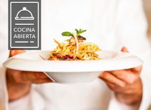 El chef presenta con maestría un plato gourmet en un impecable plato blanco con el logotipo de Cocina Abierta a la izquierda. Experimente la excelencia culinaria que captura la esencia de Badajoz en el elegante ambiente del Hotel en el Corazón Badajoz. - ForTwo - Planes para parejas