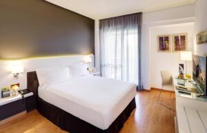 Disfrute del encanto acogedor de su moderna habitación en el Hotel en el Corazón Badajoz, equipada con una cama grande, escritorio, silla, lámparas de pared y una ventana con cortinas transparentes. El elegante suelo de madera realza este acogedor refugio. - ForTwo - Planes para parejas