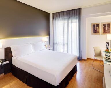 Disfrute del encanto acogedor de su moderna habitación en el Hotel en el Corazón Badajoz, equipada con una cama grande, escritorio, silla, lámparas de pared y una ventana con cortinas transparentes. El elegante suelo de madera realza este acogedor refugio. - ForTwo - Planes para parejas