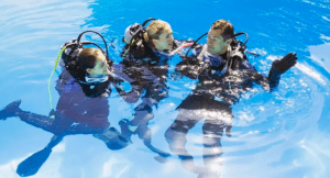 Tres buceadores con trajes de neopreno y equipo de buceo conversan durante una sesión de Iniciación Privada al Buceo en Tenerife en una piscina, y uno de ellos gesticula animadamente sobre su próxima inmersión. - ForTwo - Planes para parejas