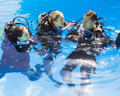 Tres buceadores con trajes de neopreno y equipo de buceo conversan durante una sesión de Iniciación Privada al Buceo en Tenerife en una piscina, y uno de ellos gesticula animadamente sobre su próxima inmersión. - ForTwo - Planes para parejas